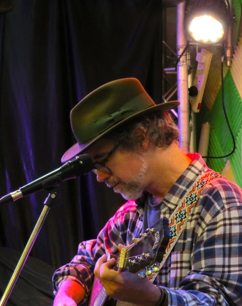 A white bearded bespectacled man in a green fedora and blue and white flannel shirt plays acoustic guitar. His head is down low in contemplation, and a microphone sits before his face though he's not currently singing into it. His guitar, reading Guild at the headstock, is partially visible at the bottom of the image, and he has a white, red, and blue embroidered strap over his shoulder.
